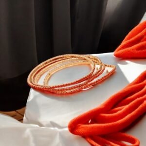 Un bracelet élastique en métal et strass en rouge corail est élégamment présenté sur un tissu blanc, accompagné d'un foulard orange. La lumière du soleil crée des ombres dynamiques qui ajoutent chaleur et dynamisme à la scène, avec le style d'accumulation tendance chic du bracelet apportant une touche d'élégance sophistiquée.