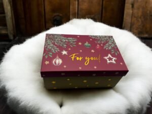 La Boîte Cadeau - Rectangulaire - Noël est un coffret cadeau décoratif au couvercle marron orné d'ornements festifs et de branches de pin, sur lequel est inscrit en jaune l'inscription « Pour toi ! ». Il est présenté posé sur un coussin blanc moelleux sur un fond en bois.