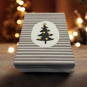 La « Boîte Cadeau - Rectangulaire - Noël - Sapin noir » arbore des rayures grises et blanches avec une étiquette ovale qui met en valeur un motif de sapin de Noël noir minimaliste. L'ambiance est encore enrichie par des lumières chaudes et floues et un fond chaleureux et festif, renforçant l'atmosphère de Noël.