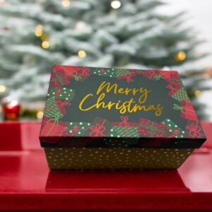 Une Boîte Cadeau Rectangulaire Noël décorée du message "Joyeux Noël" aux motifs joyeux rouges et verts repose gracieusement sur une surface rouge. En arrière-plan, un sapin de Noël flou et scintillant doucement renforce l'atmosphère festive de Noël.