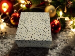 Une Boîte Cadeau Rectangulaire - Noël - Des pois gris et noirs reposent sur une surface enneigée, entourée de décorations de Noël comme des boules rouges et dorées et des lumières festives, ajoutant une touche de magie de Noël.