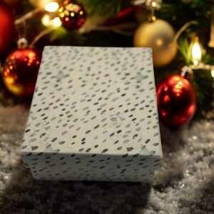 Une Boîte Cadeau Rectangulaire - Noël - Des pois gris et noirs reposent sur une surface enneigée, entourée de décorations de Noël comme des boules rouges et dorées et des lumières festives, ajoutant une touche de magie de Noël.