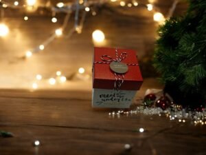 La Boîte-cadeau carrée - Noël - Rouge-blanc, ornée d'une étiquette « Joyeux Noël » et nouée avec de la ficelle rouge et blanche, trône près d'un sapin de Noël vert orné de quelques boules. Des guirlandes lumineuses chaleureuses et un fond en bois rehaussent l'ambiance chaleureuse.