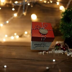 La Boîte-cadeau carrée - Noël - Rouge-blanc, ornée d'une étiquette « Joyeux Noël » et nouée avec de la ficelle rouge et blanche, trône près d'un sapin de Noël vert orné de quelques boules. Des guirlandes lumineuses chaleureuses et un fond en bois rehaussent l'ambiance chaleureuse.