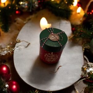 Une bougie festive verte et rouge avec un couvercle est posée sur une table entourée de décorations de Noël, notamment des boules rouges et dorées, de la verdure, des lumières chaleureuses et une Boîte-cadeau ronde - Noël - Rouge à proximité, créant une ambiance de Noël chaleureuse.