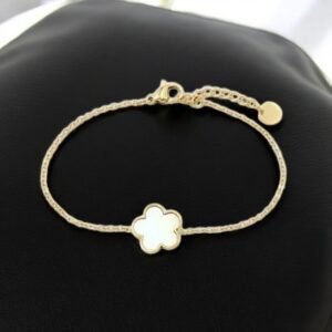 Un délicat bracelet en or avec un motif à maillons et une breloque en forme de fleur au centre, reposant sur une surface en cuir noir. Une petite étiquette circulaire est attachée près du fermoir.