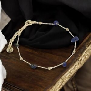 Un délicat bracelet en or avec de petites pierres bleues rondes et des accents de perles est élégamment présenté sur une surface en bois, entouré de tissu noir et blanc.