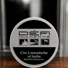 Une boîte ronde de cire à moustache et à barbe « Défi pour Homme », posée sur une surface en bois. L'étiquette comporte des icônes de soins et du texte en français et en anglais. L'arrière-plan est flou.