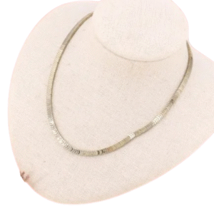 Le Collier perles flèches en hématite et acier présente de fines perles plates aux tons neutres, exposé sur un présentoir beige.