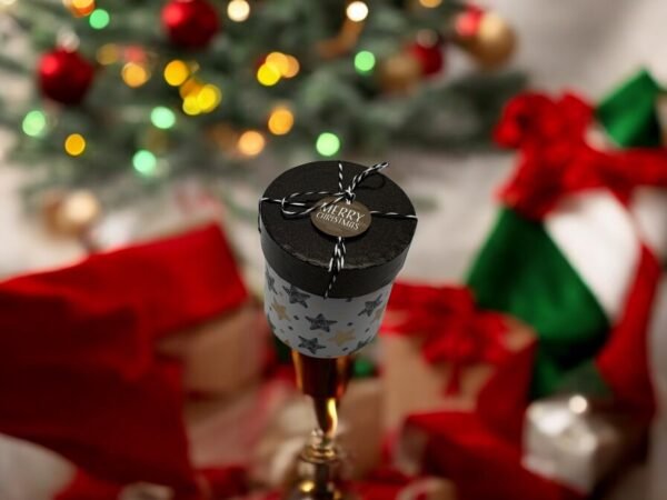 Au premier plan, une petite **Boîte-cadeau ronde-Noël-Étoiles** pend avec une étiquette « Joyeux Noël ». À l'arrière-plan, un sapin de Noël scintille de lumières, entouré d'une multitude de cadeaux emballés en rouge et vert qui renforcent l'ambiance chaleureuse des fêtes.