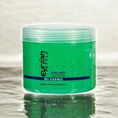 Un pot de 500 ml de gel coiffant vert étiqueté « Gel Flexible - Tenue Forte - Eyrian Style » est posé sur une surface réfléchissante, offrant une tenue « Tenue Forte ». Le fond flou renforce son attrait, le rendant idéal pour celles qui recherchent une coiffure flexible avec une tenue forte dans le style Eyrian.