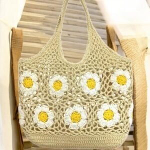 Un sac crocheté avec un motif de marguerites est suspendu à un crochet. Le sac présente six marguerites blanches et jaunes sur un fond beige et possède deux sangles. L'arrière-plan montre des planches de bois et du tissu flous.