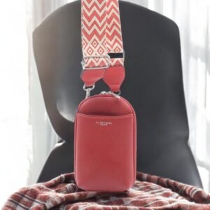 Un petit sac en cuir rouge avec une bandoulière à motif zigzag repose sur une surface en tissu rayé, rappelant le smartphone Pochette Flora&Co en similicuir marron métallisé. La pièce floue et lumineuse et la chaise sombre en arrière-plan renforcent son charme sophistiqué.