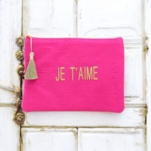 Pochette rose vif avec les mots "JE T'AIME" en lettres dorées. Le sac est doté d'une fermeture éclair à pompon doré et se présente sur un fond en bois blanc rustique.