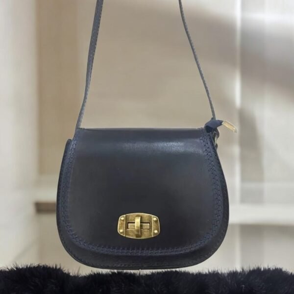 Un petit sac bandoulière en cuir foncé avec un fermoir doré est suspendu sur un fond neutre. Le sac a un design simple avec des coutures visibles sur les bords.