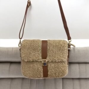 Un petit sac à main beige à la texture moelleuse avec une bandoulière et des accents en cuir marron. Il est exposé sur un canapé plissé gris clair.