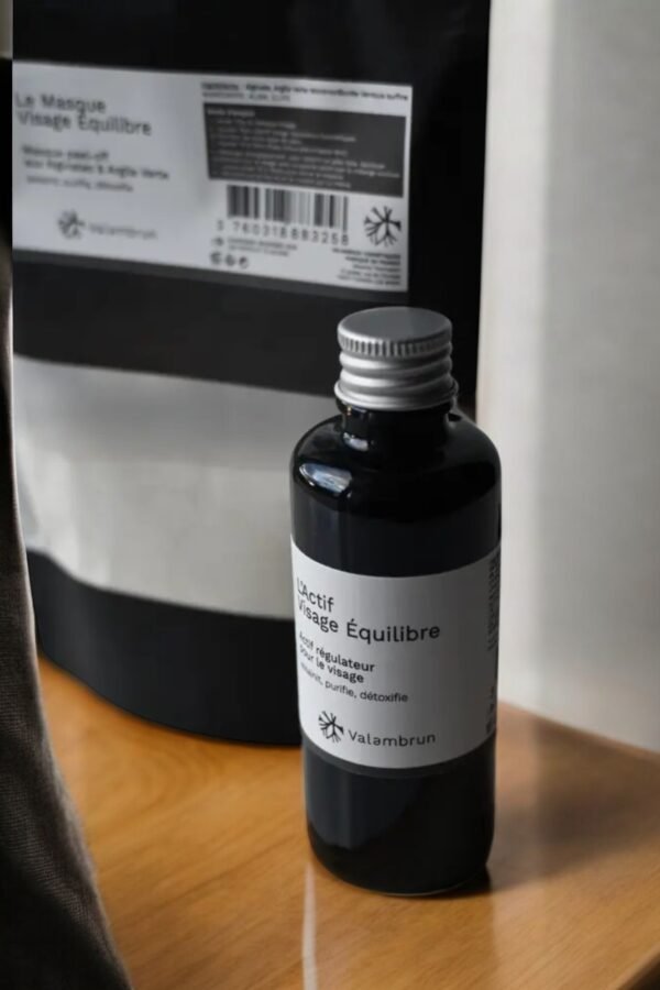 Un flacon noir étiqueté « Masque Peel Off Équilibre » de Saly Cosmétiques est posé sur une surface en bois. Derrière lui, une pochette assortie au même motif repose à côté d'un rideau blanc transparent, apportant une touche d'élégance.