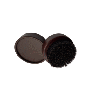 Une boîte de cirage ronde ouverte présente la Brosse à barbe Défi pour Homme posée dessus, ses poils foncés se détachant sur le fond blanc.