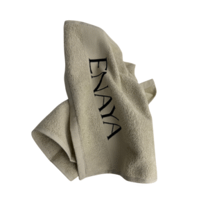 La Serviette visage Enaya, beige avec "ENAYA" brodé en noir, est partiellement pliée et légèrement froissée, présentée sur un fond blanc.