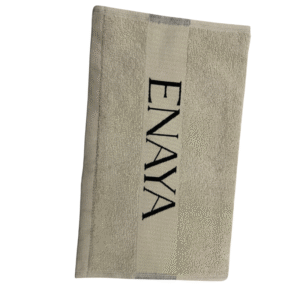 Une Serviette visage Enaya beige repose à plat sur un fond blanc, présentant une bande centrale verticale avec « ENAYA » brodé en noir.