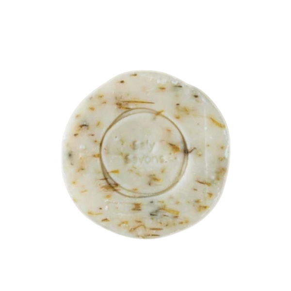 Le savon Sésame et Agrumes-120g-Saly Cosmétiques est rond et blanc cassé, avec des taches d'herbes et un motif de cercle en relief en son centre, représenté sur un fond blanc.