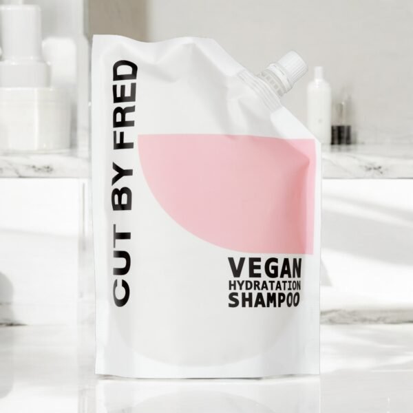 Une pochette blanche et rose « Recharge Shampooing Hydratant-Vegan-540ml-Cut by Fred » est posée sur une surface de salle de bain en marbre blanc avec un élégant flacon pompe blanc et des contenants flous en arrière-plan.