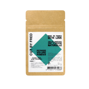 Une pochette marron étiquetée « CUT BY FRED » contient le shampooing sec Recharge Poudre Volume Vegan (10 g). L'emballage, orné de motifs géométriques verts et d'un texte bilingue noir, met en avant les ingrédients naturels et indique qu'il est fabriqué en France.