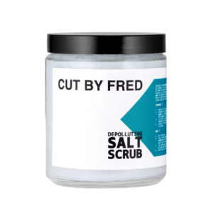 Un pot transparent avec un couvercle noir étiqueté « Gommage lavant-Depolluting Salt Scrub-300g-Cut by Fred » en blanc et bleu sarcelle contient un gommage léger conçu pour une exfoliation douce des cheveux.