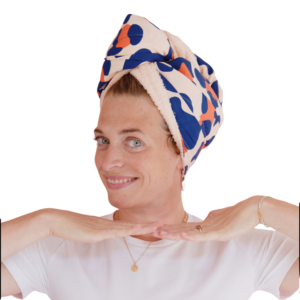 Une personne sourit, coiffée d'un turban à motifs géométriques By Fred bleu, rose et orange. Elle lève les mains près de son visage pour montrer la douceur obtenue grâce au shampoing et gel douche Recharge Baby. Elle porte une chemise blanche et des bijoux en or sur un fond uni.