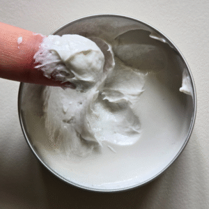 Un doigt prélève l'épaisse Gomme coiffante blanche Flex-ultra maniable-Défi pour Homme (75 ml) dans un récipient métallique rond et ouvert sur une surface légère.