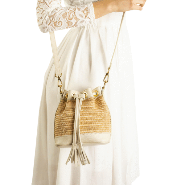 Une femme en robe de dentelle blanche tient le sac Raphia et cuir véritable - Bandoulière réglable-Vanille avec cordon et glands, porté en bandoulière. L'image présente un fond blanc uni.