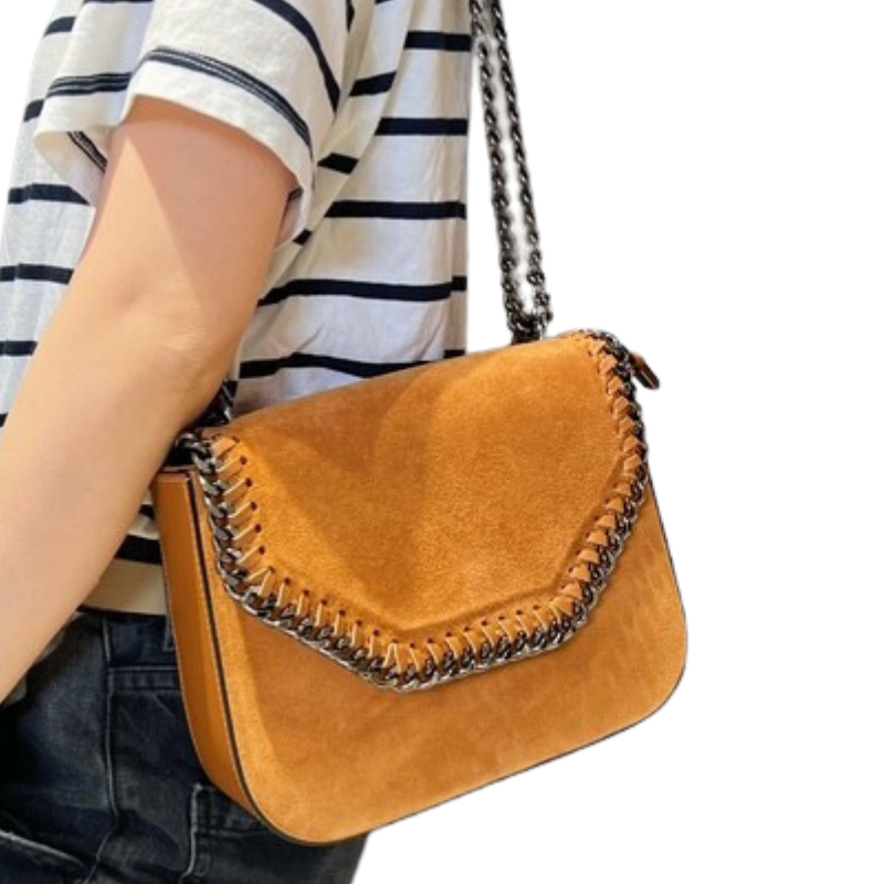 Une personne vêtue d'une chemise rayée à manches courtes et d'un jean porte le Sac à bandoulière en daim avec détail chaîne-Camel, un sac à bandoulière en daim fauve doté d'un détail chaîne audacieux sur le rabat et d'une sangle en chaîne métallique, suspendu à son côté.