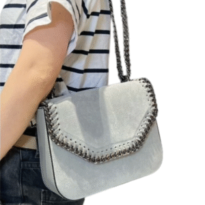 Une personne portant une chemise à rayures noires et blanches et un pantalon noir porte le Sac à bandoulière en daim avec détail chaîne-Gris, un sac à bandoulière gris clair doté d'une sangle en chaîne et d'un détail décoratif en chaîne sur le bord du rabat.