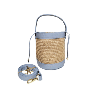 Le Sac à main bandoulière raphia garni cuir-Bleu Ciel est un sac cylindrique en raphia avec une garniture en cuir bleu clair, une poignée supérieure assortie et une bandoulière détachable avec des ferrures dorées, présenté sur un fond blanc.