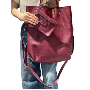 Une personne en jean bleu et haut beige tient le Sac à main en cuir irisé et cuir grainé-Bordeaux, un grand fourre-tout en cuir marron avec pochette amovible et sangle réglable.