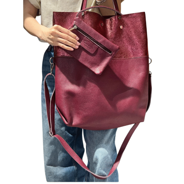 Une personne en jean bleu et haut beige tient le Sac à main en cuir irisé et cuir grainé-Bordeaux, un grand fourre-tout en cuir marron avec pochette amovible et sangle réglable.