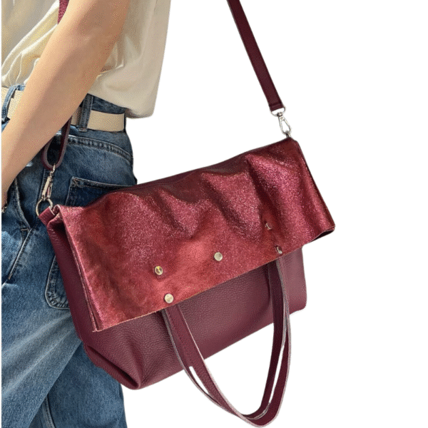 Une personne en jean et haut blanc porte le Sac à main en cuir irisé et cuir grainé-Bordeaux, un sac chic de couleur bordeaux avec un rabat brillant, des clous métalliques, des bandoulières et des poignées sur fond blanc.