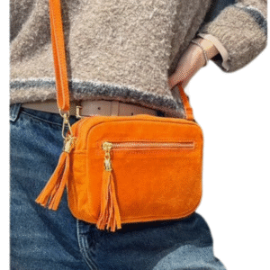 Une personne vêtue d'un jean bleu et d'un pull beige texturé porte le Sac caméra à bandoulière en daim, un petit sac rectangulaire en daim orange avec une fermeture éclair à pompon et des ferrures dorées.