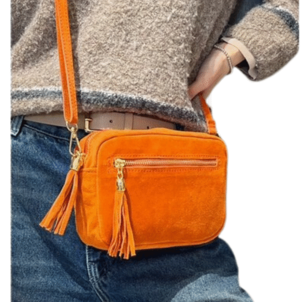 Une personne vêtue d'un jean bleu et d'un pull beige texturé porte le Sac caméra à bandoulière en daim, un petit sac rectangulaire en daim orange avec une fermeture éclair à pompon et des ferrures dorées.