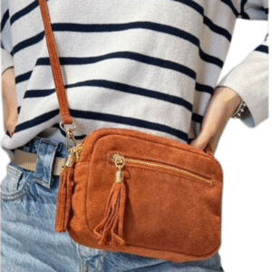Une personne vêtue d'un pull rayé blanc et marine et d'un jean bleu porte le Sac caméra à bandoulière en daim- Rouille, un petit sac rectangulaire à bandoulière en daim marron avec une fermeture éclair dorée, des ferrures dorées et un pompon décoratif.