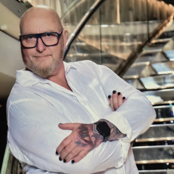Un homme chauve avec des lunettes, une courte barbe et du vernis à ongles noir pose les bras croisés, montrant un tatouage sur son avant-bras. Il porte une chemise blanche et se tient devant un escalier moderne - un look audacieux à propos de Hair Passion.