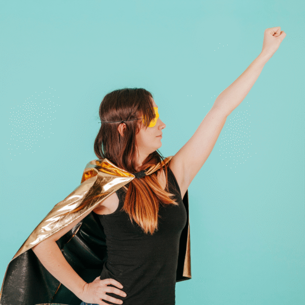 Une femme portant une cape dorée et un masque jaune prend la pose d'une super-héroïne, levant un bras avec assurance sur un fond bleu clair - un visuel qui capture l'esprit d'autonomisation A propos de Hair Passion.