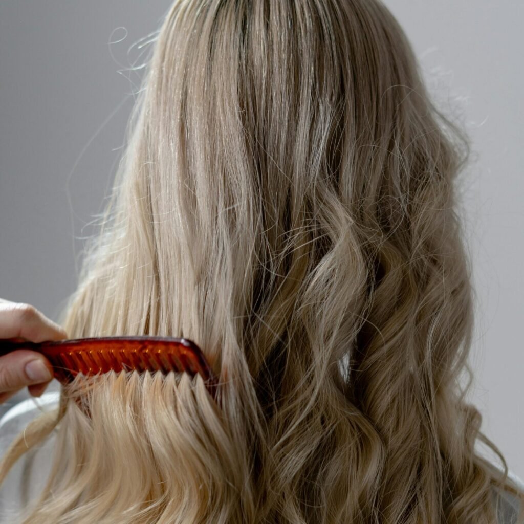 Une personne aux longs cheveux blonds ondulés se fait coiffer avec un peigne à dents larges brun-rouge. Capturée de dos, l'image met en valeur l'accueil hair passion pour un démêlage en douceur et des résultats impeccables.