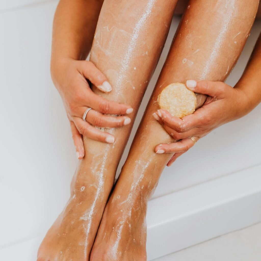 Une personne assise sur le bord d'une baignoire exfolie le bas de ses jambes à l'aide d'un gommage rond, montrant une peau humide et brillante et des ongles d'orteils bien entretenus - l'accueil parfait pour toute routine de soins personnels ou de passion capillaire.