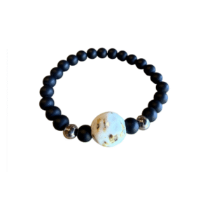 Le Bracelet en billes volcaniques / pierre bleue se compose de perles noires mates, de deux perles argentées brillantes et d'une grande perle blanche avec des marques brunâtres, le tout sur fond blanc.