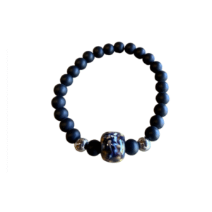 Le Bracelet en billes volcaniques /pierre bleue se compose de perles volcaniques noires mates, de deux petits accents métalliques argentés et d'une perle décorative plus grande avec un motif bleu et blanc, sur fond blanc.