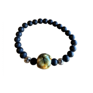 Le Bracelet en billes volcaniques /pierre bleue se compose de perles rondes noires mates, de deux perles argentées brillantes et d'une grande perle centrale multicolore polie, sur fond blanc.