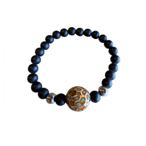 Le Bracelet en billes volcaniques /pierre bleue se compose de perles volcaniques noires mates, de deux accents argentés brillants et d'une perle centrale avec un motif en nid d'abeille doré et turquoise, sur fond blanc.