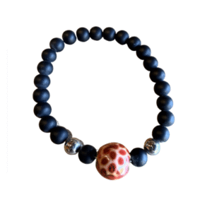 Le Bracelet en billes volcaniques /pierre bleue se compose de perles volcaniques noires mates, de deux accents argentés brillants et d'une pièce centrale unique à pois rouges et beiges, sur fond blanc.