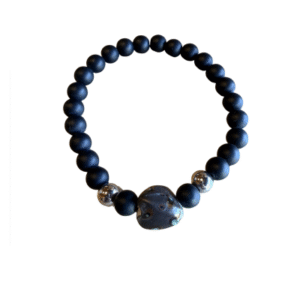 Le Bracelet en billes volcaniques /pierre bleue se compose de perles volcaniques noires mates, de deux accents argentés brillants et d'une pièce centrale unique en pierre bleue, le tout présenté sur un fond blanc.